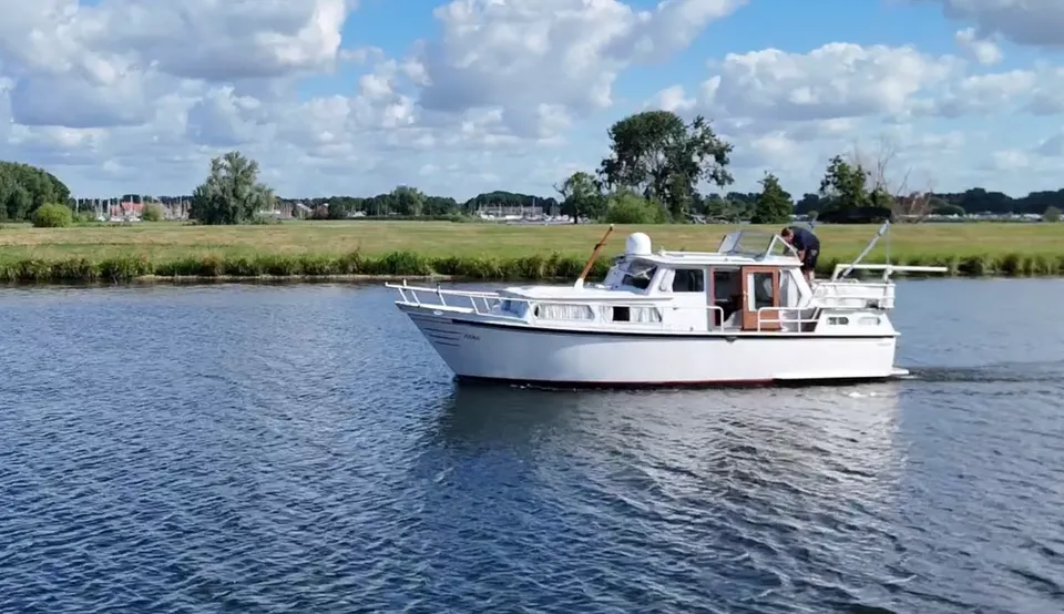 Een witte motorboot vaart op blauw water met een groene oever en bomen op de achtergrond onder een bewolkte hemel.