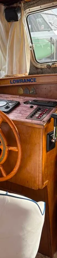 De foto toont het interieur van een boot met een houten stuurwiel, bedieningspaneel, witte stoel en ramen met uitzicht op…