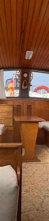 De foto toont het interieur van een boot met houten lambrisering, een witte stoel, banken, een tafel en ramen met gordijnen.