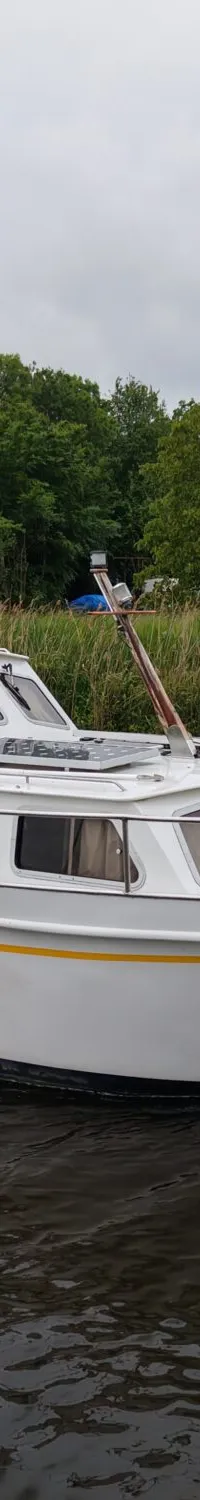 Een witte motorboot met de naam "Ruel" vaart op een waterweg met groene oevers onder een bewolkte lucht.