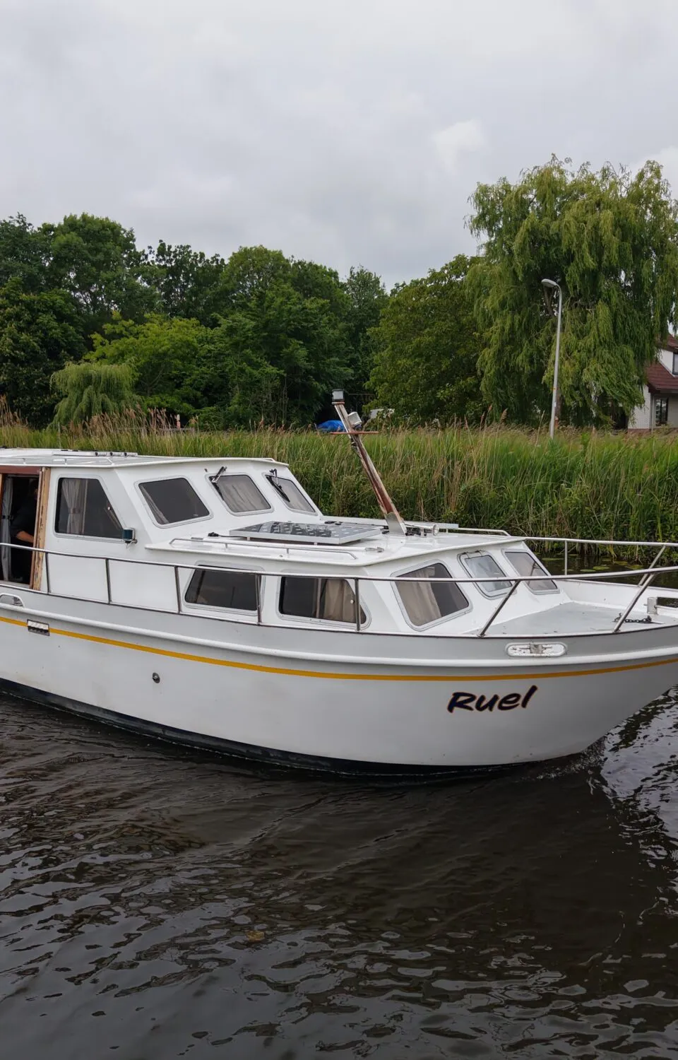 Een witte motorboot met de naam "Ruel" vaart op een waterweg met groene oevers onder een bewolkte lucht.