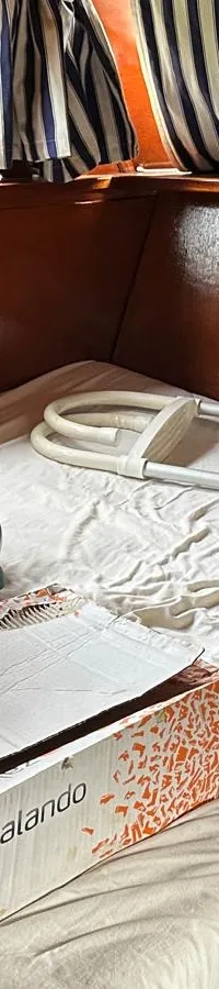 De foto toont een kajuit met houten wanden, een bed met wit laken, een Zalando-doos en een opvouwbare trolley.