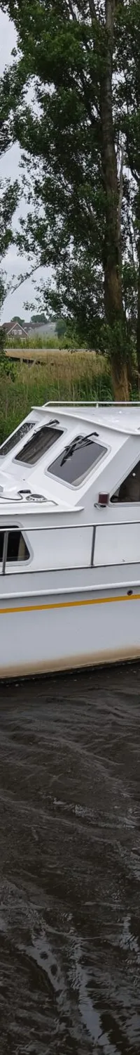 Een witte motorboot met de naam "Ruel" op de boeg, drijvend op donker water, omringd door groen en bomen.