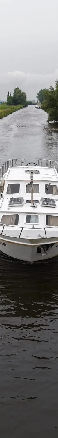 Een witte motorboot vaart op een kanaal met groene oevers onder een bewolkte hemel.
