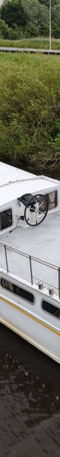 Een witte motorboot met een geel accent op de romp vaart op een kanaal langs een groene oever met een weg en gebouwen op de…