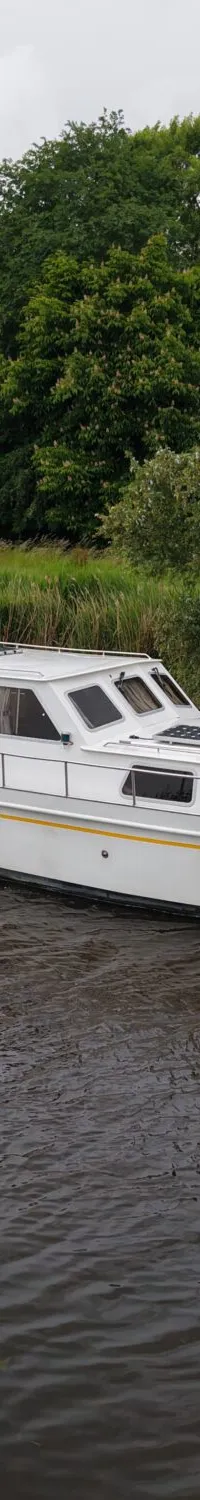 Een witte motorboot met de naam "Ruel" vaart op een kanaal met groene oevers en bomen onder een bewolkte hemel.