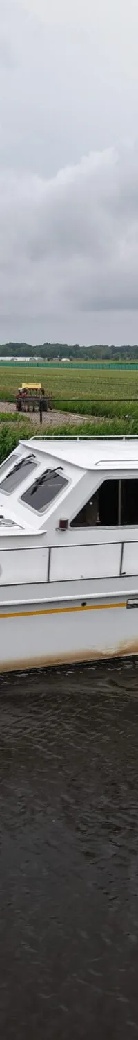 Een witte motorboot met de naam "Ruel" vaart op een waterweg, met een groene schuur en velden op de achtergrond.