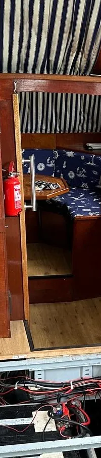 De foto toont het interieur van een boot met een houten stuurwiel, een open vloerluik met bedrading en een televisie.