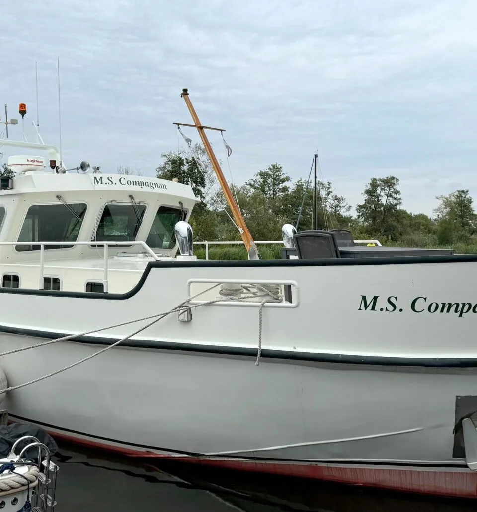 Een witte boot met de naam "M.S. Compagnon" ligt afgemeerd aan een houten steiger.