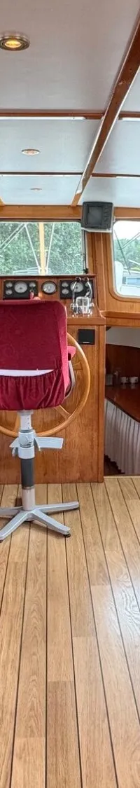 De foto toont het interieur van een boot met houten lambrisering, rode zitbanken en krukken, een stuurstand en ramen met…
