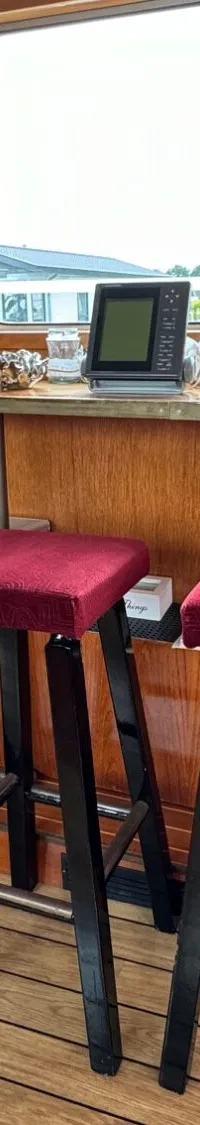 De foto toont het interieur van een boot met houten wanden, een houten vloer, drie barkrukken met rode zittingen en een…
