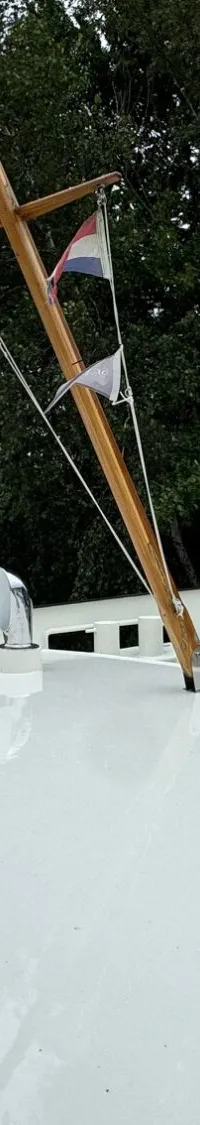Een wit dek met een houten mast met twee Nederlandse vlaggen, een tafel met stoelen en een luik.