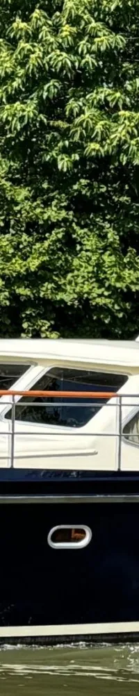 Een donkerblauwe motorboot met een crèmekleurige opbouw vaart op een groene waterweg, omringd door bomen.