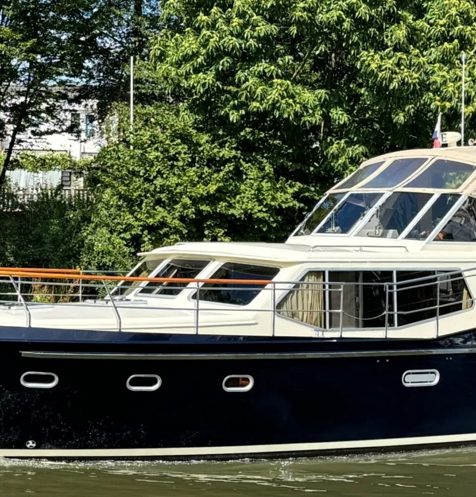 Een donkerblauwe motorboot met een crèmekleurige opbouw vaart op een groene waterweg, omringd door bomen.