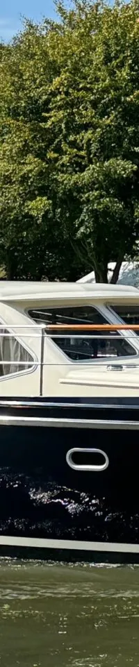 Een donkerblauwe motorboot met een crèmekleurige opbouw vaart op een waterweg met bomen en gebouwen op de achtergrond.