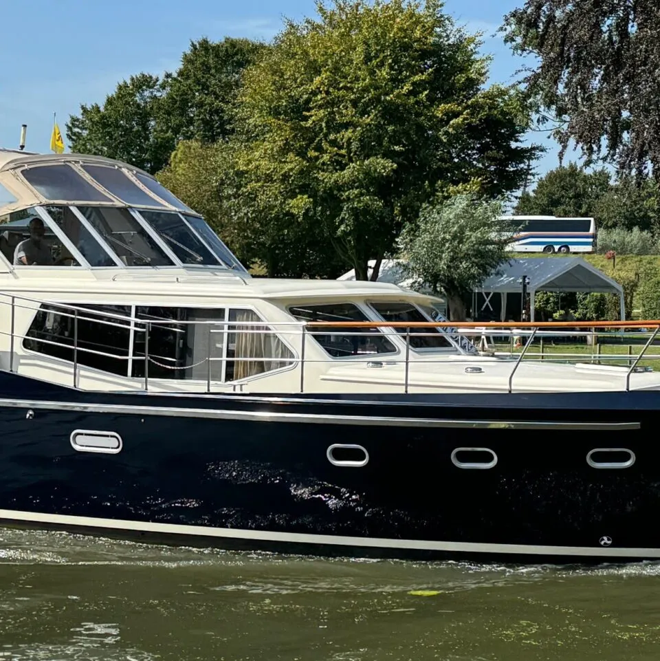 Een donkerblauwe motorboot met een crèmekleurige opbouw vaart op een waterweg met bomen en gebouwen op de achtergrond.