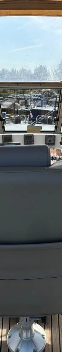 Een stuurhut met een grijze stuurstoel, houten vloer en een uitzicht op een jachthaven.
