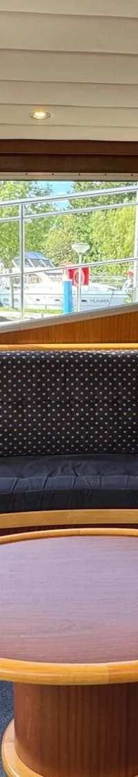 De foto toont een interieur van een boot met een U-vormige bank, een ronde tafel, ramen met gele gordijnen en een kleine…