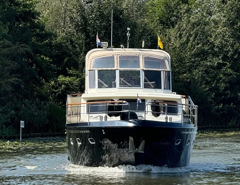 Een donkerblauwe motorboot met een lichte opbouw vaart op een waterweg met bomen op de achtergrond.