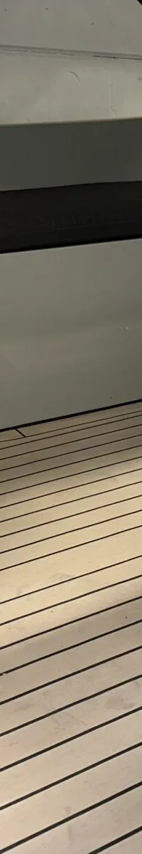 De foto toont het interieur van een boot met een lichtgekleurde vloer met zwarte naden, grijze banken met donkere kussens…
