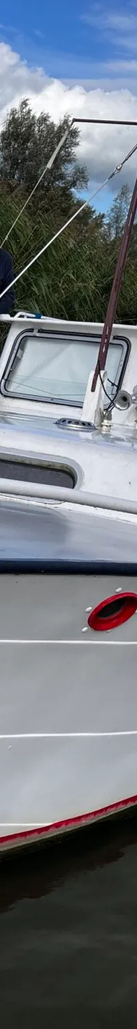 Een witte motorboot met een wapenschild op de boeg ligt aangemeerd aan een steiger, met een man aan boord.