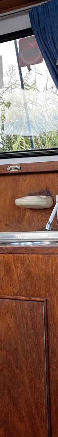 De foto toont een houten interieur van een boot met een keukenblok, ramen met gordijnen en een blauwe zitting.