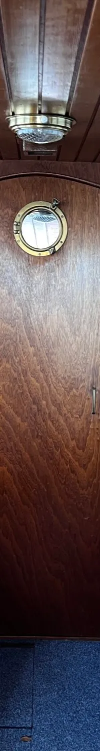 De foto toont het interieur van een kajuit met houten lambrisering, twee banken met gele kussens en een patrijspoort in de…