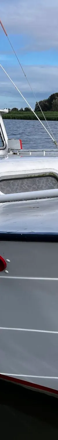 Een witte motorboot met de letters "JM" op de kajuit, ligt afgemeerd aan een steiger met een rivier op de achtergrond.