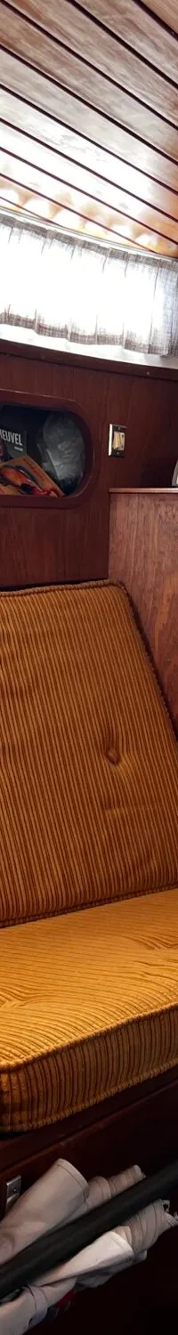 De foto toont een houten kajuit met twee banken met okergele ribfluwelen kussens, ramen met gordijnen en een patrijspoort.