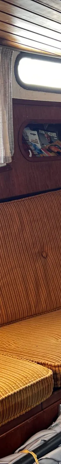 De foto toont het interieur van een boot met een houten plafond en wanden, een oranje gestreepte bank, ramen en een…