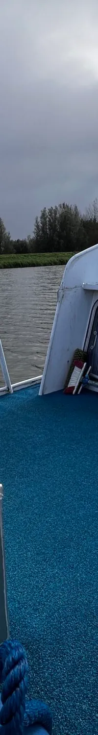 Een boot met een blauw dek, een stuurwiel en een aluminium opbergkist, varend op een rivier onder een bewolkte lucht.
