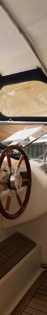 Een interieur van een boot met een stuurstand, een houten stuurwiel, een witte stoel en een instrumentenpaneel.