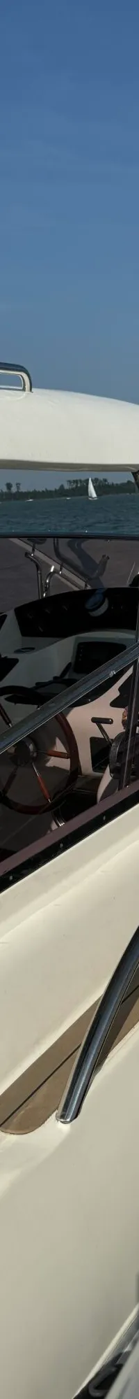 Een witte motorboot met een donkerblauwe kap en een houten stuurwiel, liggend op het water met zeilboten op de achtergrond.