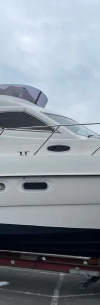Een witte motorboot met een donkerblauwe romp staat op een rode trailer op een parkeerplaats onder een bewolkte hemel.