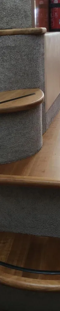 Een trap met houten treden en grijze vloerbedekking leidt naar beneden, met een houten vloer en een bank zichtbaar.