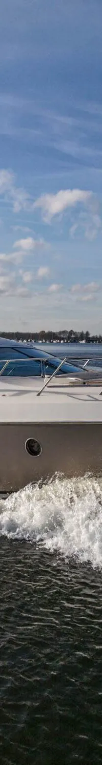 Een grijs-wit motorjacht vaart door het water, met een Nederlandse vlag aan de boeg, en creëert boeg- en hekgolven.