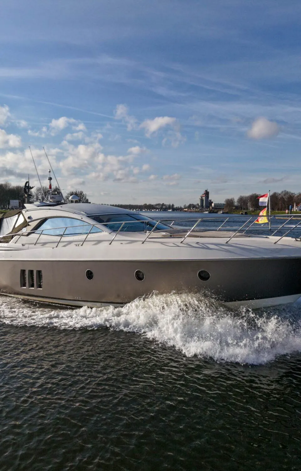 Een grijs-wit motorjacht vaart door het water, met een Nederlandse vlag aan de boeg, en creëert boeg- en hekgolven.