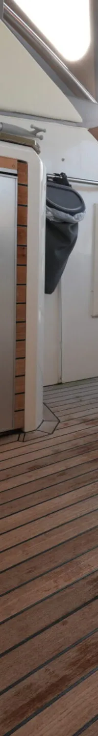 De foto toont een bootinterieur met een houten dek, een roestvrijstalen koelkast, een bedieningspaneel en zitplaatsen.