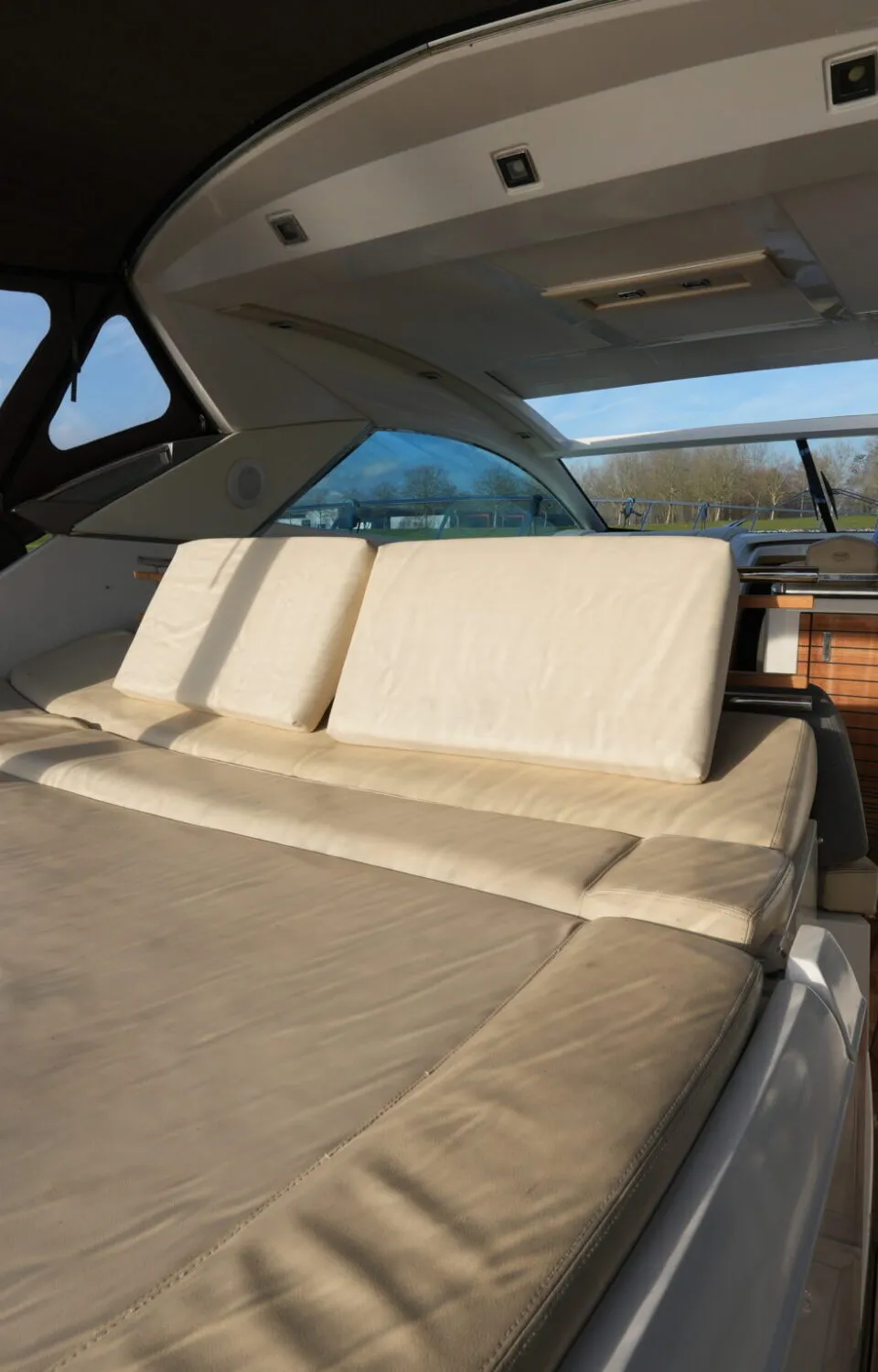 De foto toont een deel van een bootinterieur met beige kussens, een houten vloer en een stuurstand op de achtergrond.