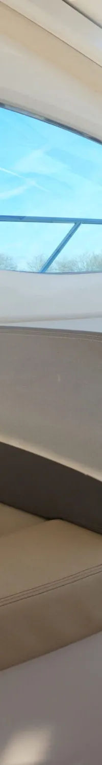 Een interieurfoto van een boot toont beige zitkussens, een houten tafel en vloer, en grote ramen met uitzicht op de blauwe…