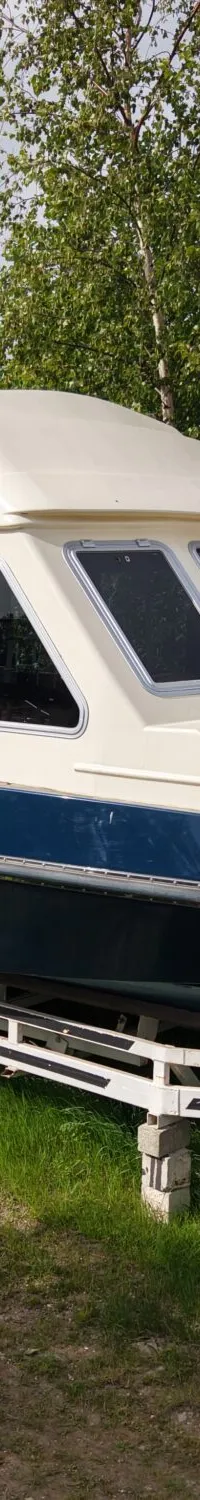 Een blauw-witte kajuitboot staat op een witte trailer met dubbele as, geparkeerd op gras naast een golfplaten schuur.