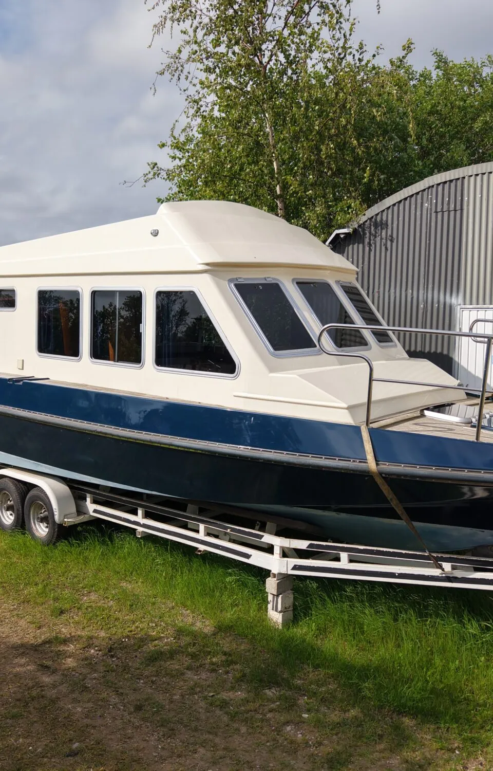 Een blauw-witte kajuitboot staat op een witte trailer met dubbele as, geparkeerd op gras naast een golfplaten schuur.