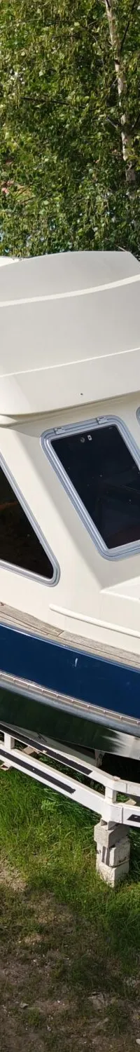 Een blauw-witte kajuitboot staat op een trailer op een grasveld, met op de achtergrond containers en een loods.