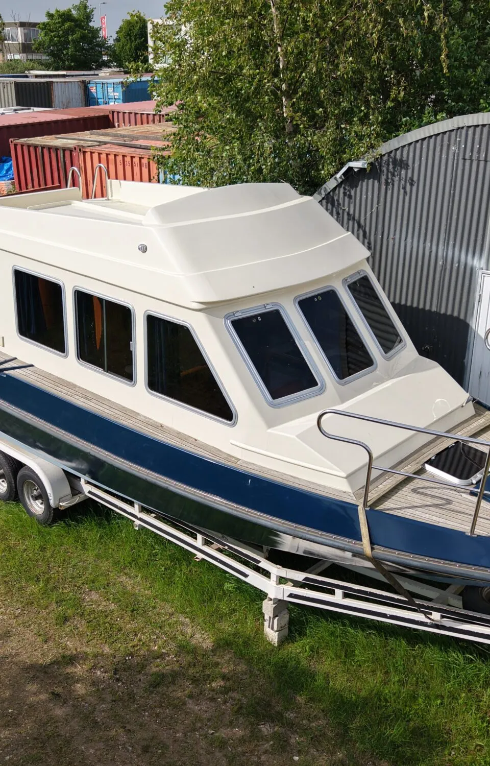 Een blauw-witte kajuitboot staat op een trailer op een grasveld, met op de achtergrond containers en een loods.