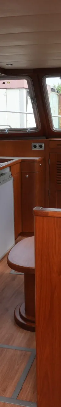 De foto toont het interieur van een boot met houten afwerking, een kombuis, zithoek en stuurstand.