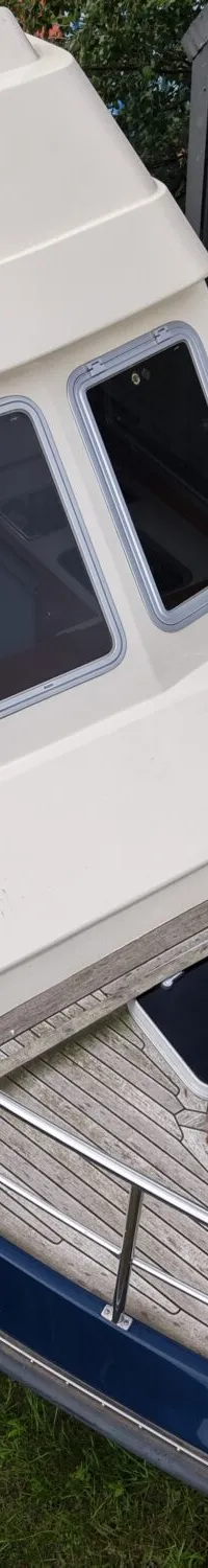 Een blauw-witte boot met houten dek staat op een trailer naast een golfplaten schuur met een witte deur.