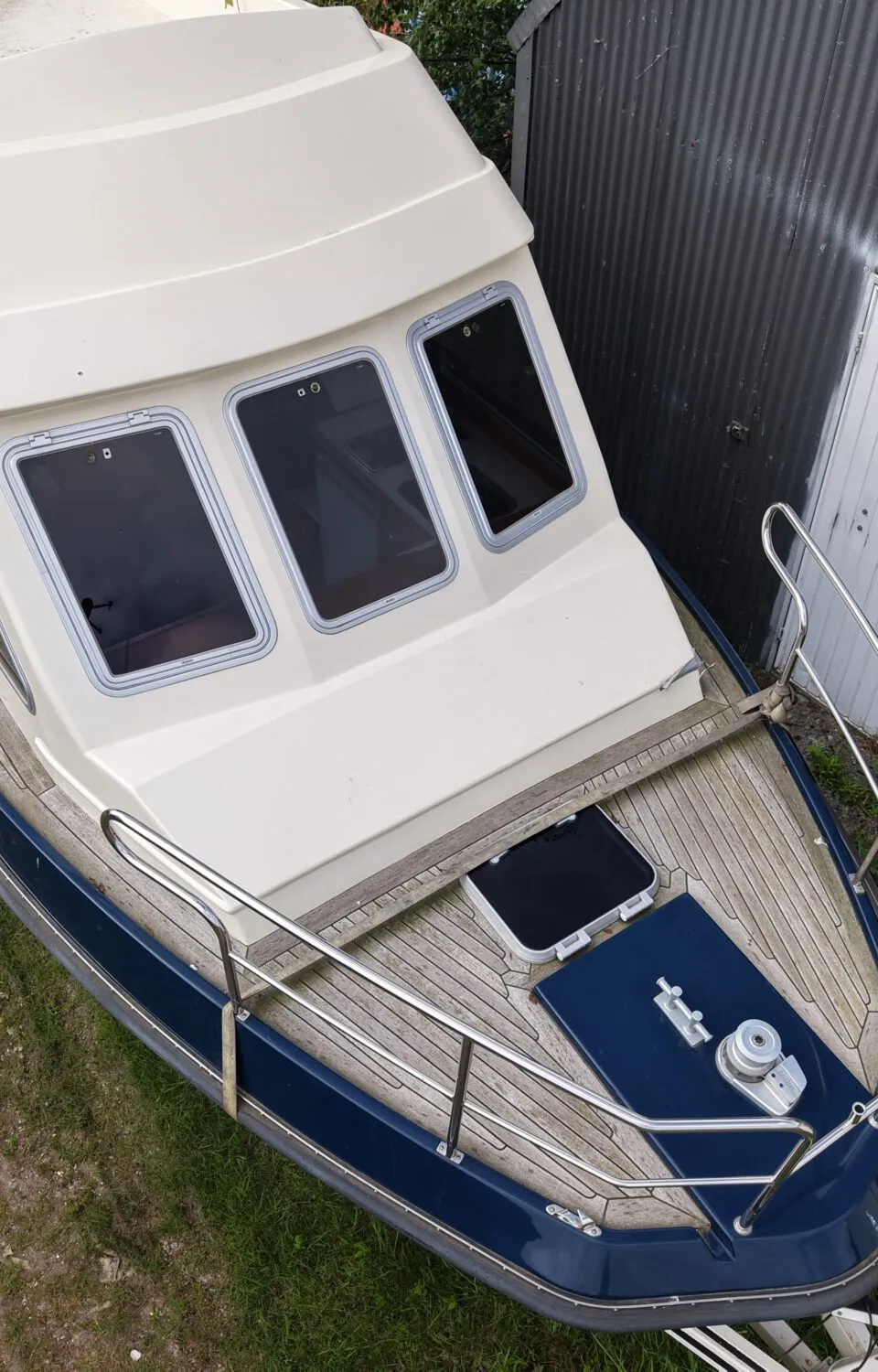 Een blauw-witte boot met houten dek staat op een trailer naast een golfplaten schuur met een witte deur.