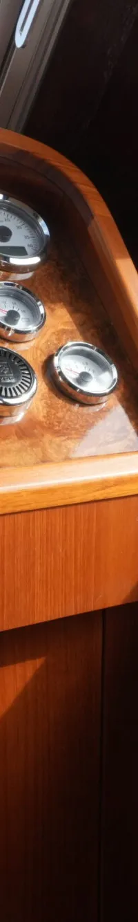 Een houten dashboard met diverse meters en schakelaars, met een raam dat uitkijkt op een blauw zeil en de horizon.