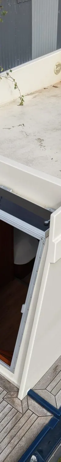 Een crèmekleurige boot met een houten dek en een open deur naar de kajuit, staand op gras naast struiken en gebouwen.