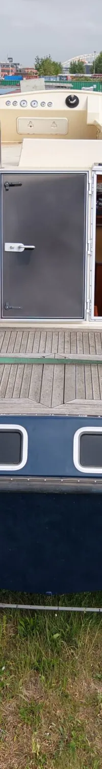 Een blauwe boot met een houten dek en een crèmekleurige opbouw staat op het gras naast een waterweg.